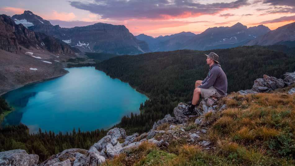 An Adventist man communing with God on Saturday Sabbath, sitting on rocks near a lake next to a forest, looking at the mountain range across.