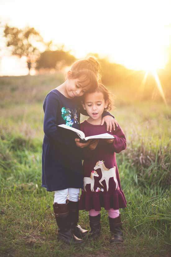 An older sister embraces her younger sister as they stand outside in the sun and read the Bible