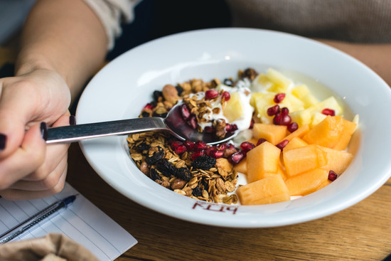 A hand holding a spoon and eating granola with fresh berries and cantaloupe, components of a healthy lifestyle